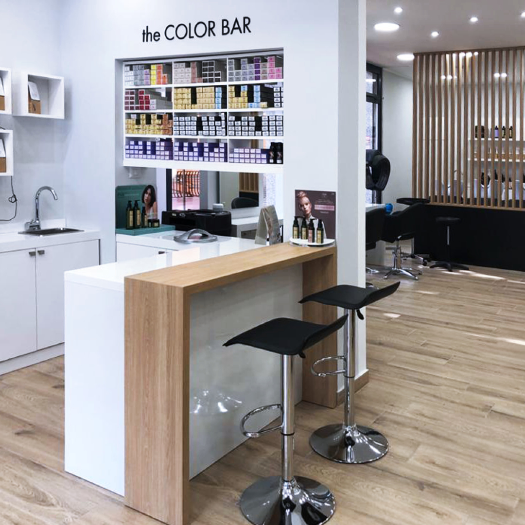 Immagine del bancone in legno, con davanti due sgabelli neri e dietro tutti i colori per capelli